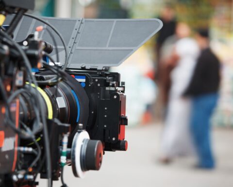 A video camera is in the foreground of the frame and is pointed towards a group of people, who are out of focus in the background.