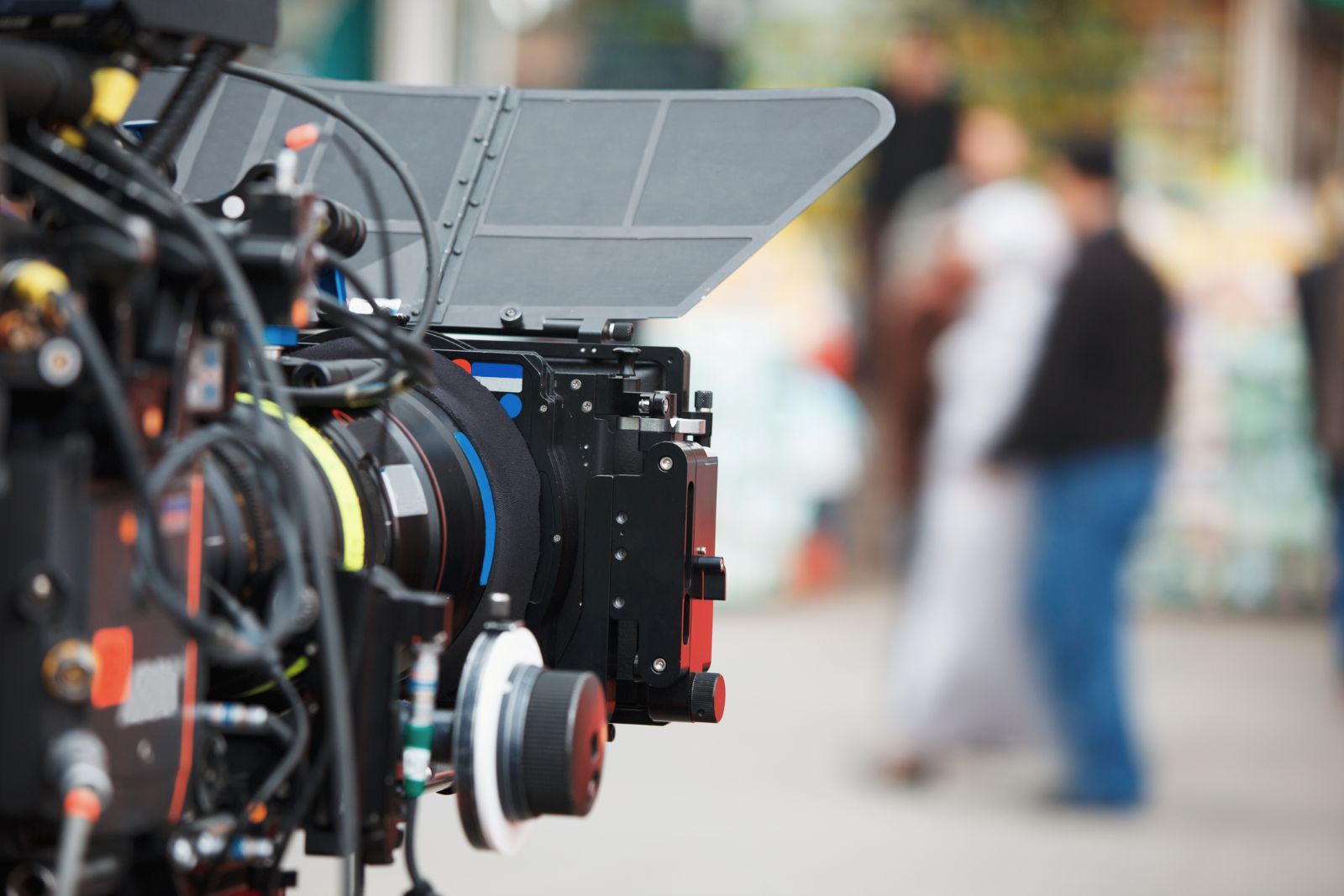 A video camera is in the foreground of the frame and is pointed towards a group of people, who are out of focus in the background.