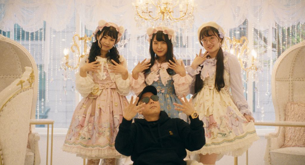 Chef Masaki Saito sits like a gangster in a black jacket and shades, surrounded by young women in frilly outfits.