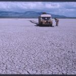 A beige Land Rover sits in the distance in a barren mud-cracked field.