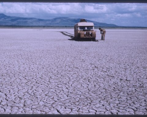 A beige Land Rover sits in the distance in a barren mud-cracked field.