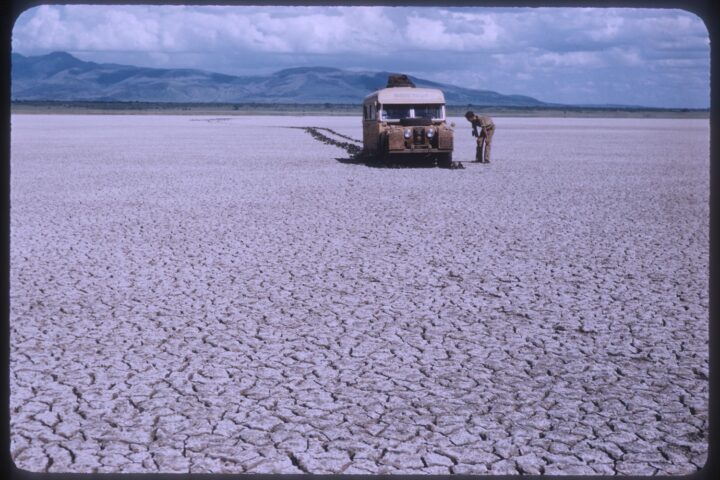 A beige Land Rover sits in the distance in a barren mud-cracked field.