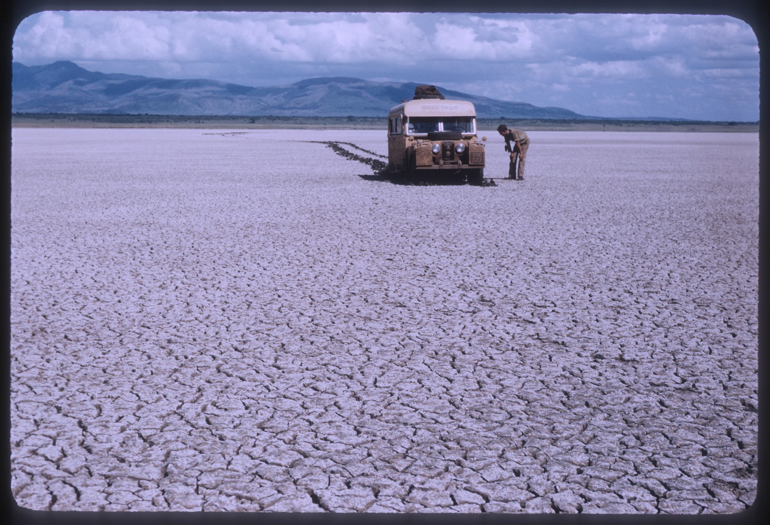 A beige Land Rover sits in the distance in a barren mud-cracked field.