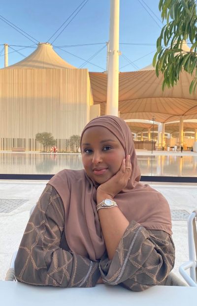 Ruun Nuur is a Black Muslim woman. She is pictured sitting down outside wearing a pink hijab. She is resting her head on her hand, looking at the camera, and smiling.