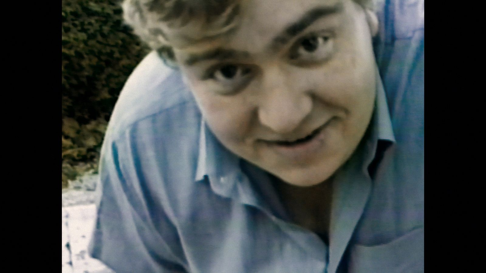Actor John Candy appears in close-up in a VHS family home movie. He is bending over and looking directly at the camera.