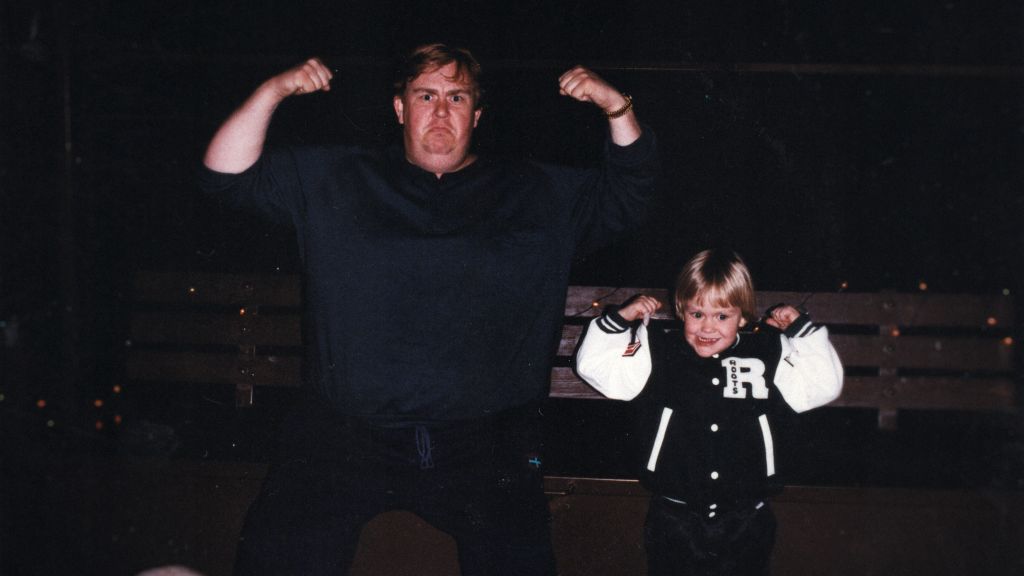 An old family photo of actor John Candy and his son Chris. They are standing with their arms raised, against a black background.