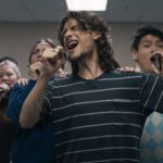 A diverse group of university age students rehearses a song performance. They are standing in a v formation with their hands raised holding water bottles and cell phones as microphones.