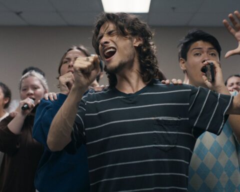 A diverse group of university age students rehearses a song performance. They are standing in a v formation with their hands raised holding water bottles and cell phones as microphones.