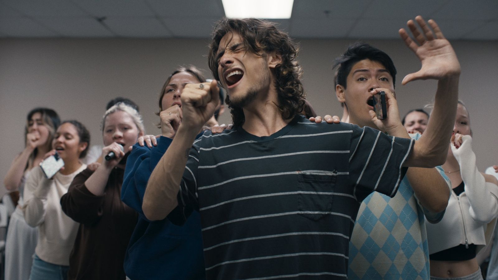 A diverse group of university age students rehearses a song performance. They are standing in a v formation with their hands raised holding water bottles and cell phones as microphones.