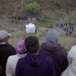 A group of people atop a hill look towards a crowd at ground level in the distance. There is a white tent in the middle of the field.