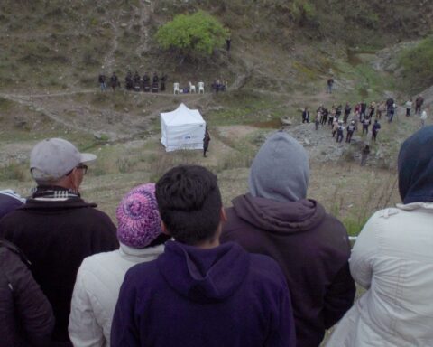 A group of people atop a hill look towards a crowd at ground level in the distance. There is a white tent in the middle of the field.