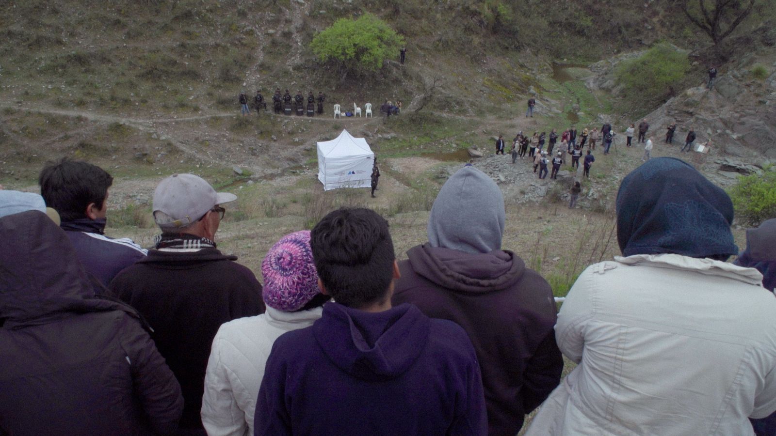 A group of people atop a hill look towards a crowd at ground level in the distance. There is a white tent in the middle of the field.