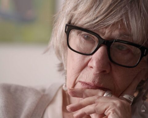 A close up portrait of an older woman with her chin on her hand, listening. She is wearing black glasses and has grey hair.