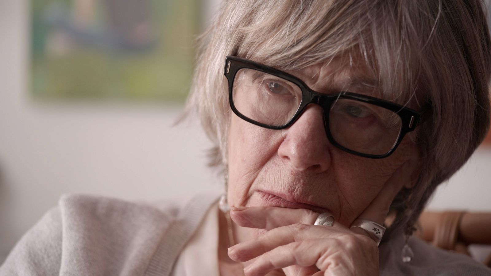 A close up portrait of an older woman with her chin on her hand, listening. She is wearing black glasses and has grey hair.