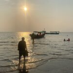 A silhouette of a man on a beach. He is where the water meets the shore, walking towards a boat in the distance.