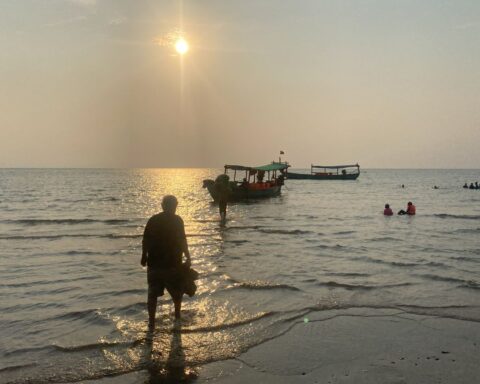 A silhouette of a man on a beach. He is where the water meets the shore, walking towards a boat in the distance.