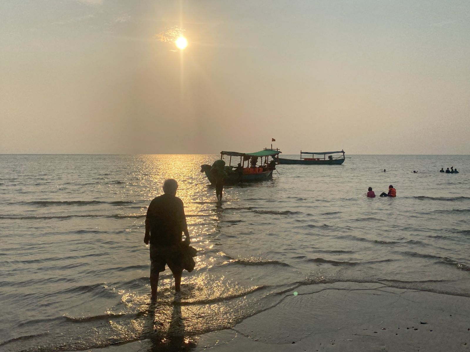 A silhouette of a man on a beach. He is where the water meets the shore, walking towards a boat in the distance.