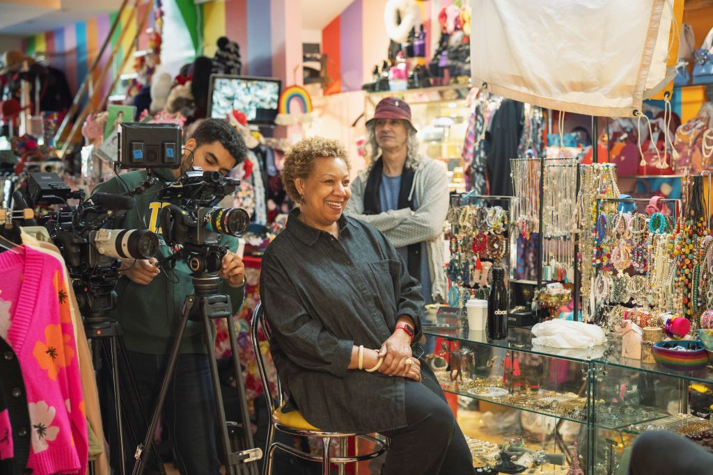 Filmmaker Alison Duke, a Black woman with short gold hair, sits in a chair conducting an interview in a clothing store. She is wearing a black shirt and black pants. A cameraperson is behind her.