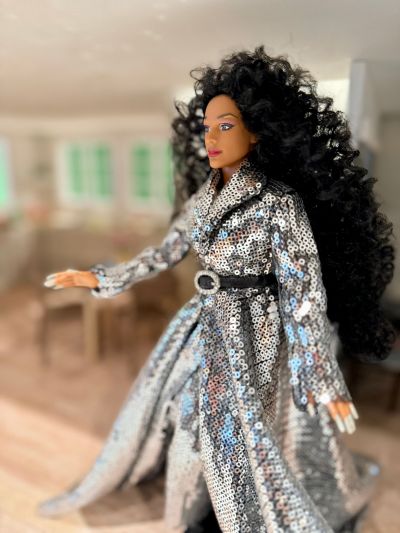 A Black doll resembling Michelle Ross is dress in a silvery coat with black belt, and flowing curly black hair.