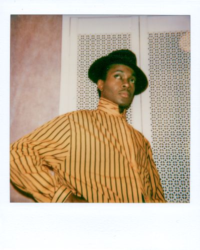 A photo of a Black man in the 1980s. He is wearing a yellow and brown striped collared shirt and a black hat. He is standing in front of white patterned doors.
