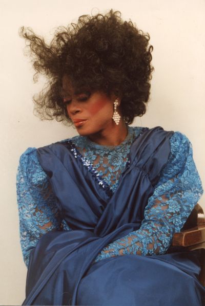 A photo of Michelle Ross. The Black drag queen is pictured in profile wearing a blue sleeved dress. Her face is turned to the left and her hair is blowing across it.