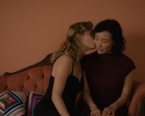 A 53-year-old Chinese woman wearing a black dress receives a kiss from a younger blonde woman. They are seated on a pink couch and the wall behind them is rose-coloured.