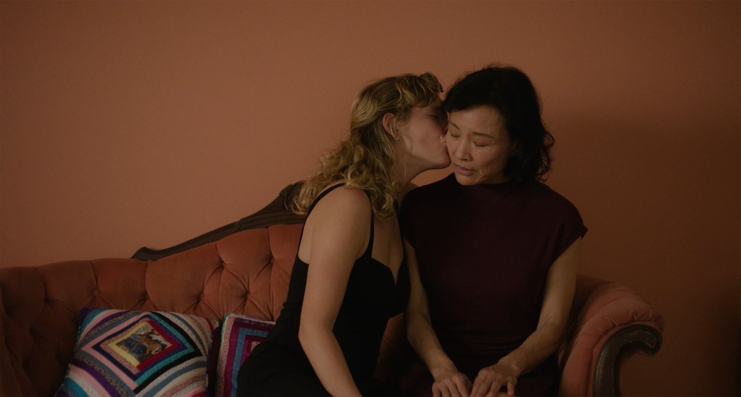 A 53-year-old Chinese woman wearing a black dress receives a kiss from a younger blonde woman. They are seated on a pink couch and the wall behind them is rose-coloured.