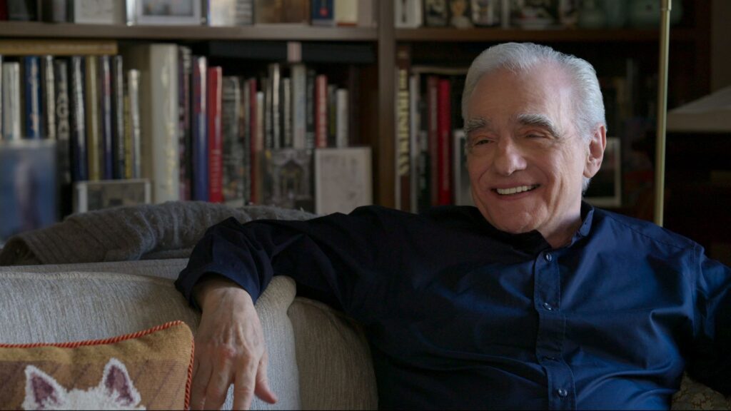Filmmaker Martin Scorsese is an Italian-American man in his 80s. He is seated on a couch, wearing a blue collared shirt, and is laughing. There is a full bookcase behind him.