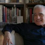 Filmmaker Martin Scorsese is an Italian-American man in his 80s. He is seated on a couch, wearing a blue collared shirt, and is laughing. There is a full bookcase behind him.