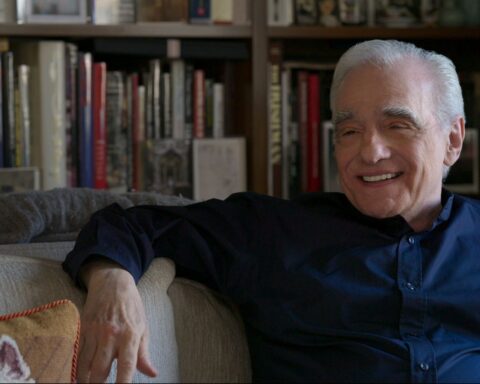 Filmmaker Martin Scorsese is an Italian-American man in his 80s. He is seated on a couch, wearing a blue collared shirt, and is laughing. There is a full bookcase behind him.
