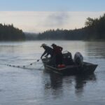 Two people stand in a motorboat on a river. They are pulling in a net from the water.