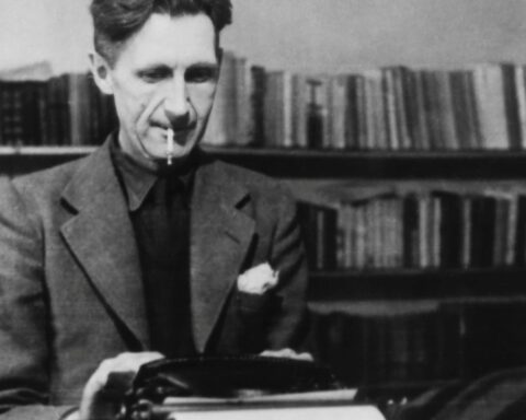 A black and white photograph of author George Orwell. He is seated at a desk, wearing a suit, and typing on a typewriting while smoking a cigarette.