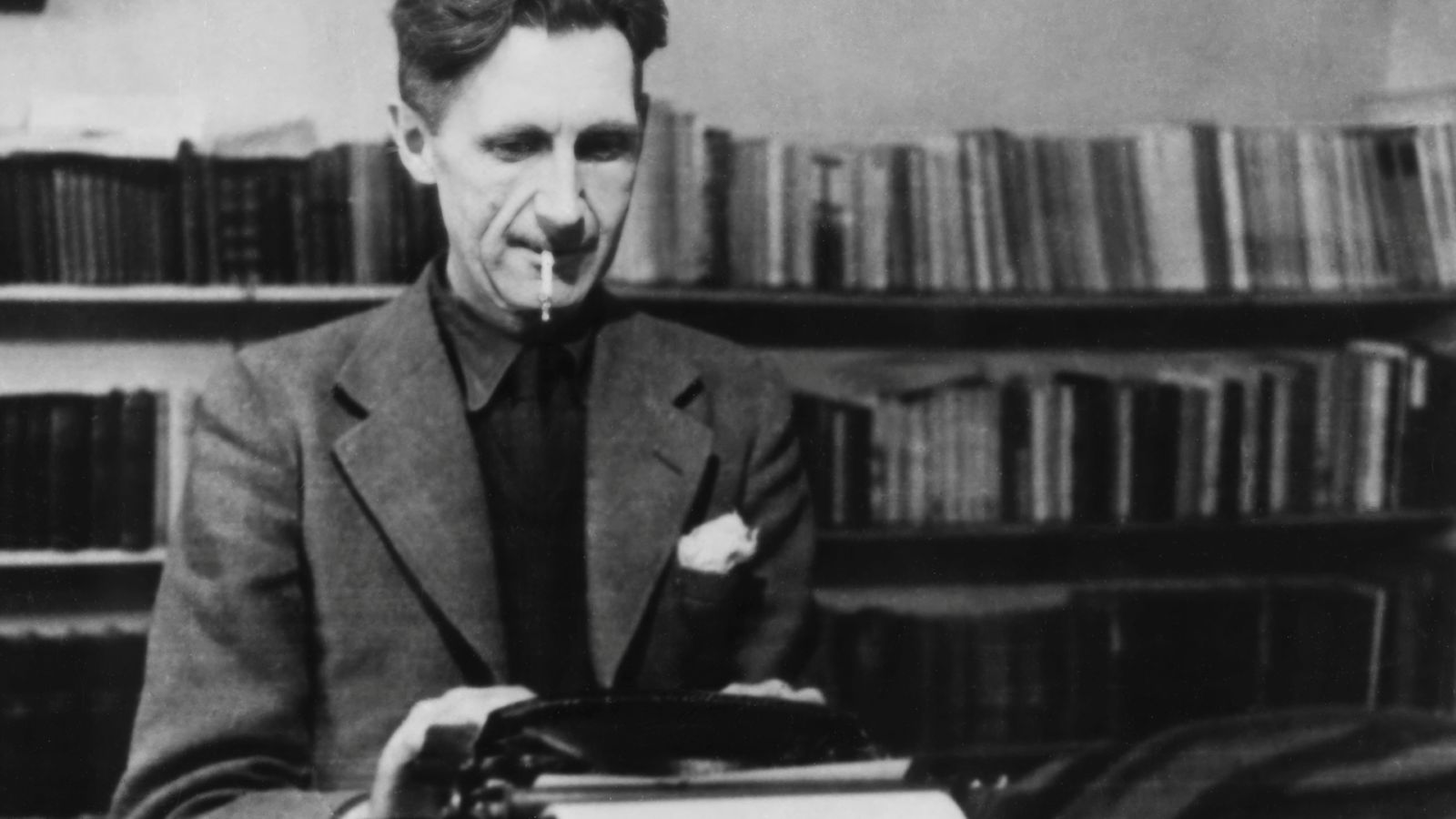 A black and white photograph of author George Orwell. He is seated at a desk, wearing a suit, and typing on a typewriting while smoking a cigarette.