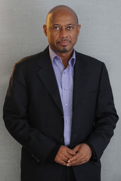 Raoul Peck is a Black man wearing a black blazeer and light blue shirt. He is pictured from the waist up, looking at the camera with his hands folded at the waist.