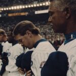 Players from the Montreal Expos stand in a line at the side of the baseball field holding their ball caps to their hearts. Their uniforms are white with thin blue stripes and blue hats. The stadium behind them is crowded.