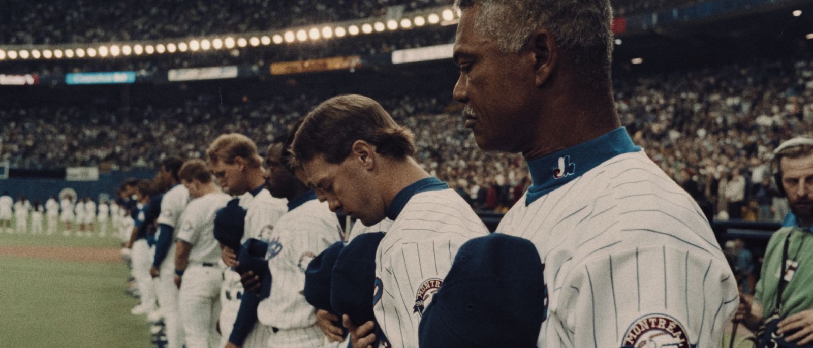 Players from the Montreal Expos stand in a line at the side of the baseball field holding their ball caps to their hearts. Their uniforms are white with thin blue stripes and blue hats. The stadium behind them is crowded.