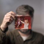 A man holds a photograph of a young girl in front of his face. His face is covered by the photo. He is wearing a brown sleeved shirt.