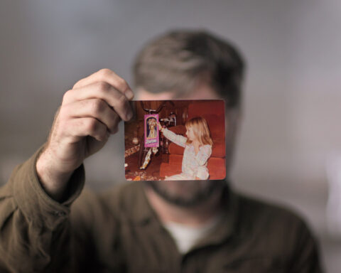 A man holds a photograph of a young girl in front of his face. His face is covered by the photo. He is wearing a brown sleeved shirt.