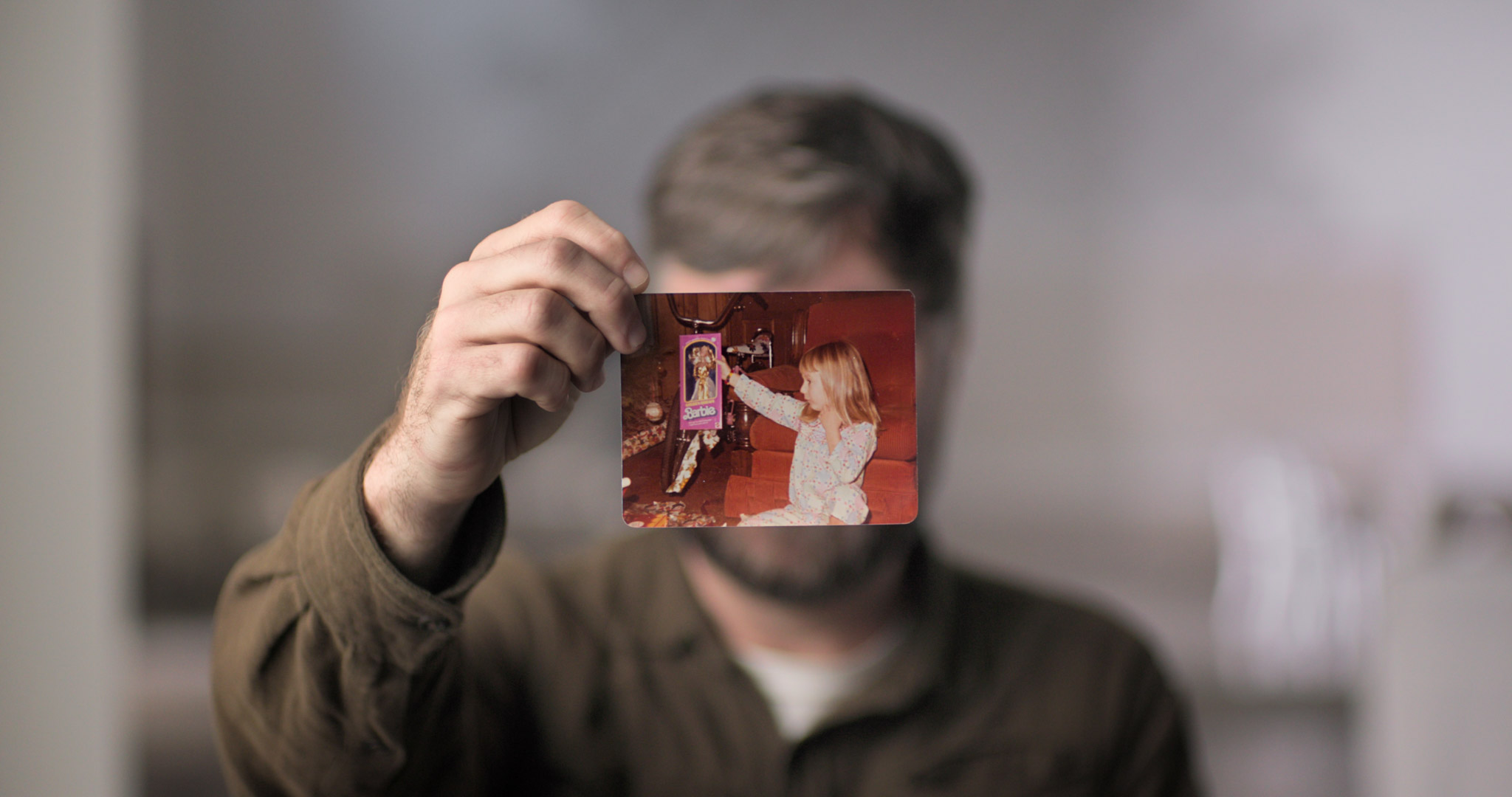 A man holds a photograph of a young girl in front of his face. His face is covered by the photo. He is wearing a brown sleeved shirt.
