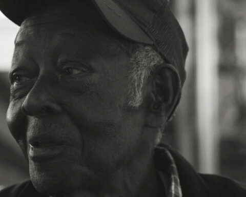A black and white photo shows an elderly Black farmer in close-up. He is smiling and looking to the left, and wearing a baseball cap.