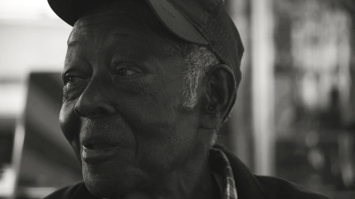 A black and white photo shows an elderly Black farmer in close-up. He is smiling and looking to the left, and wearing a baseball cap.