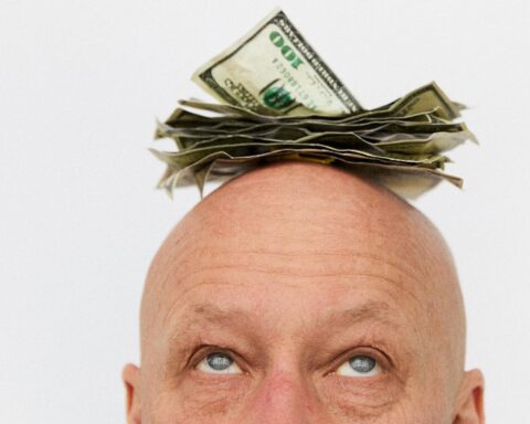 A close-up image of a bald man looking upward at a stack of money atop his head. He has blue eyes and the background is white.