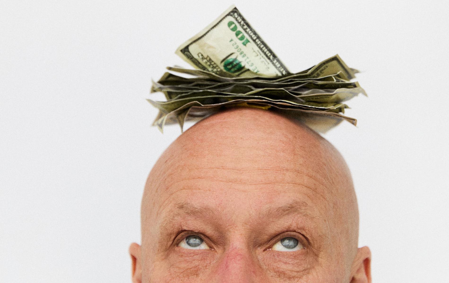 A close-up image of a bald man looking upward at a stack of money atop his head. He has blue eyes and the background is white.