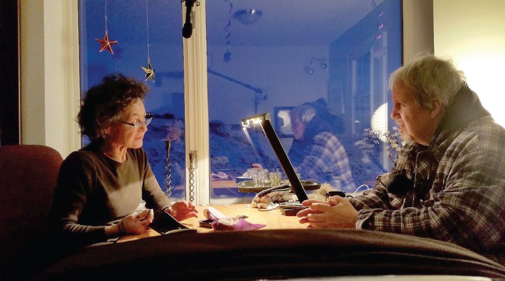An Inuk woman and a white man sit at a desk having a conversation. There is a microphone between them.