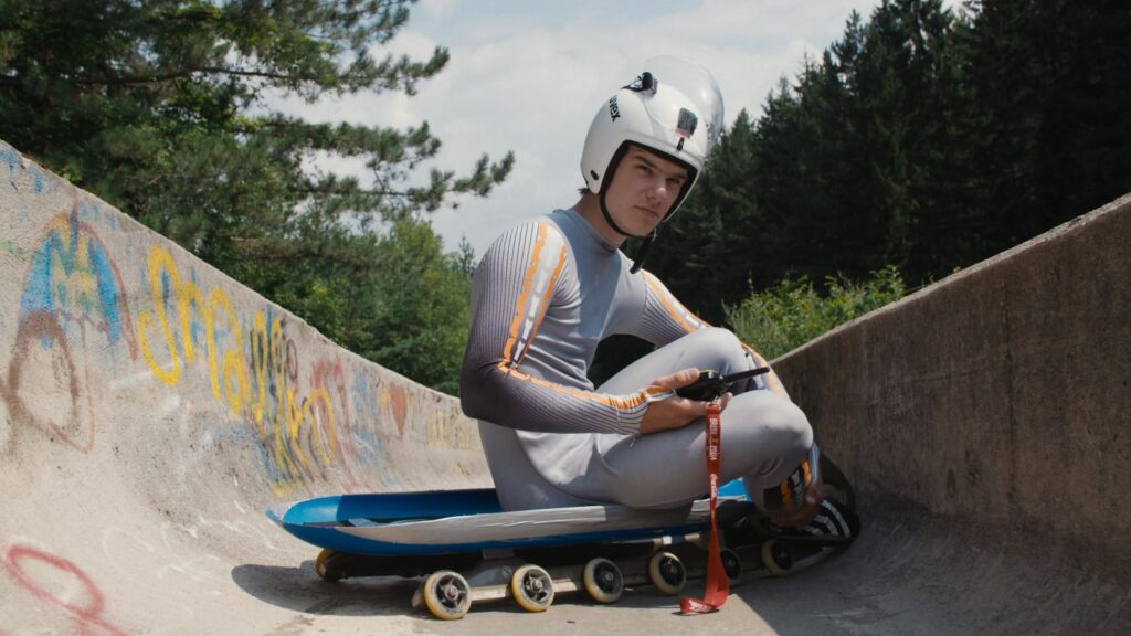 A luge athlete in his late teens/early twenties sits on a sled in a concrete luge track covered in graffiti.