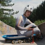 A luge athlete in his late teens/early twenties sits on a sled in a concrete luge track covered in graffiti.