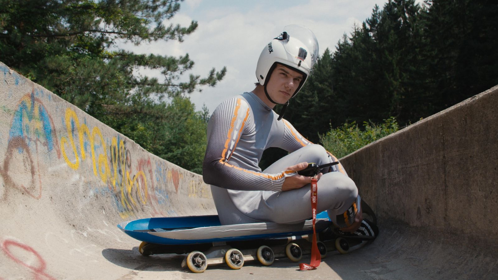 A luge athlete in his late teens/early twenties sits on a sled in a concrete luge track covered in graffiti.