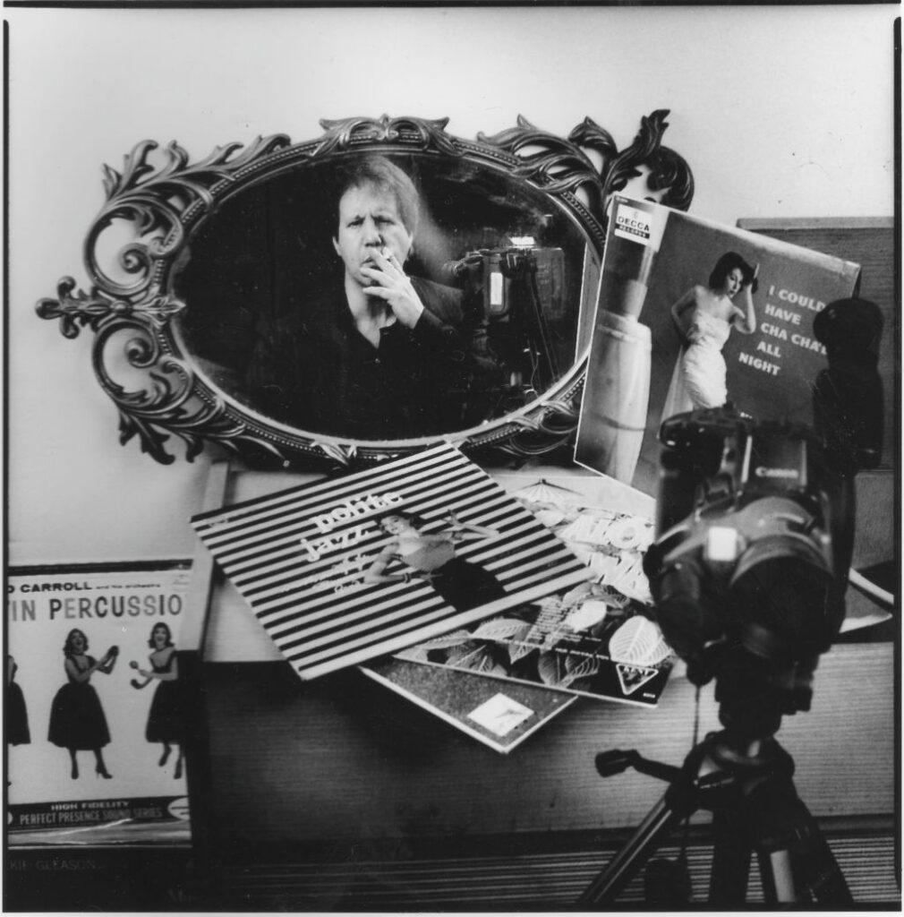 A black and white photo of a mirror propped on a dresser. There is a camera facing it and recording the reflection of a filmmaker as he smokes a cigarette.