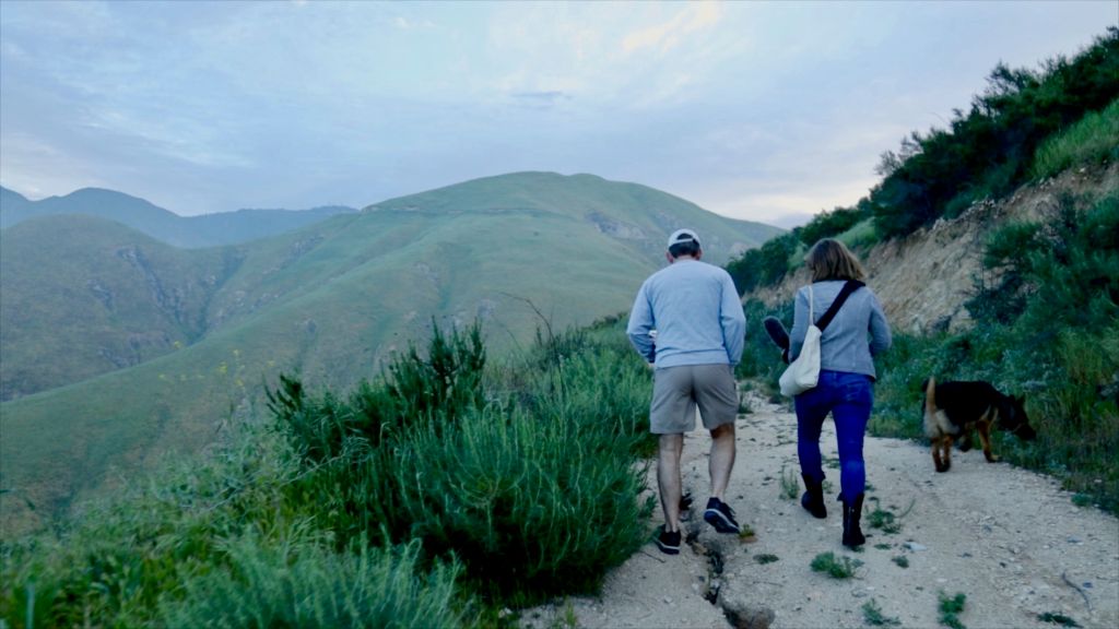 Two people walk along a path at the edge of a mountain. There are rolling green hillsides in the background.