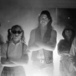A black and white photo of the band Butthole Surfers. Five people stand looking at the camera with their arms crossed. There is a lens flare in the middle of the photo.